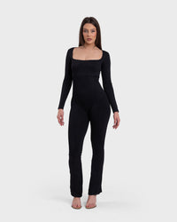 A woman modeling a long-sleeve black jumpsuit with a square neckline and flare design, standing against a white background.